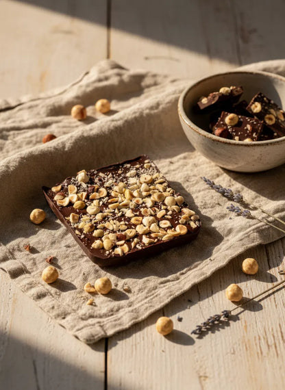 Dark chocolate bar with chopped hazelnuts on a linen cloth, beside a bowl of chocolate pieces and lavender sprigs.