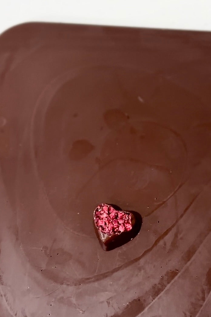 Single dark chocolate heart, topped with vibrant dried raspberries, on a smooth brown surface.