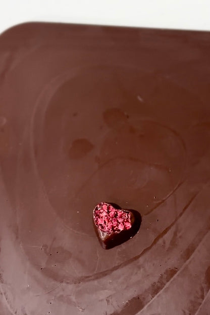 Single dark chocolate heart, topped with vibrant dried raspberries, on a smooth brown surface.