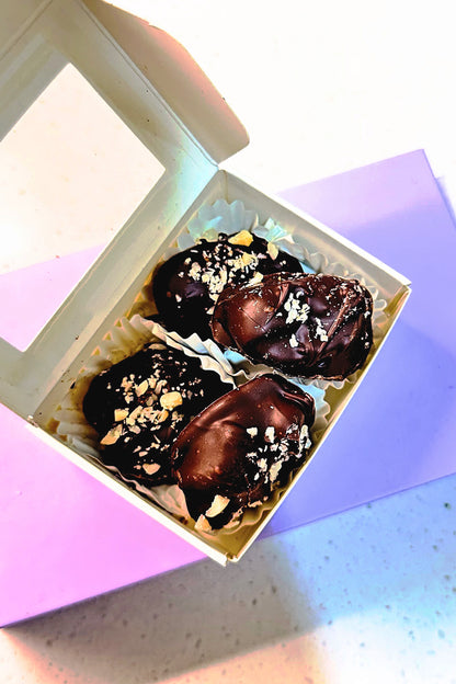 Four dark chocolate-coated prunes, sprinkled with nuts, in a white box on a purple surface.