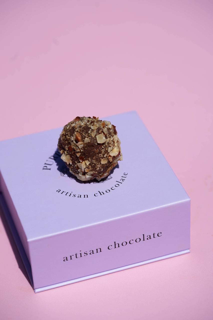 Dark chocolate truffle with chopped nuts on a light purple "Artisan Chocolate" box.