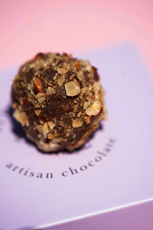 Dark chocolate truffle covered in chopped nuts, resting on a light purple box with "artisan chocolate" text.