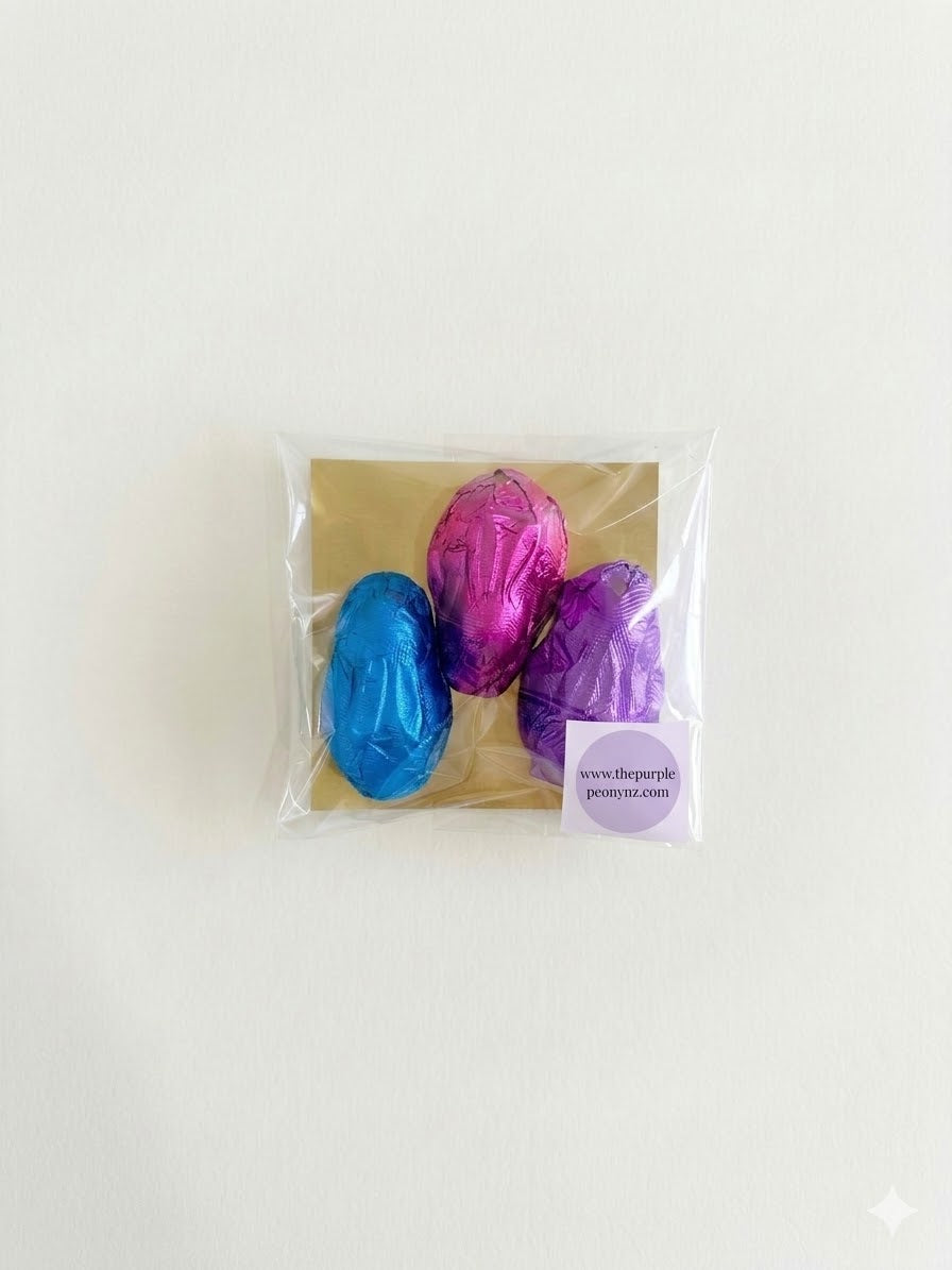 Three foil-wrapped chocolate Easter eggs in blue, pink, and purple, packaged in a clear bag with a brown backing.