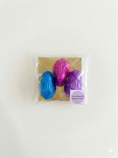 Three foil-wrapped chocolate Easter eggs in blue, pink, and purple, packaged in a clear bag with a brown backing.