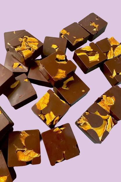 Dark chocolate Mango Bites, square pieces with visible golden dried mango chunks, scattered on a light purple surface.