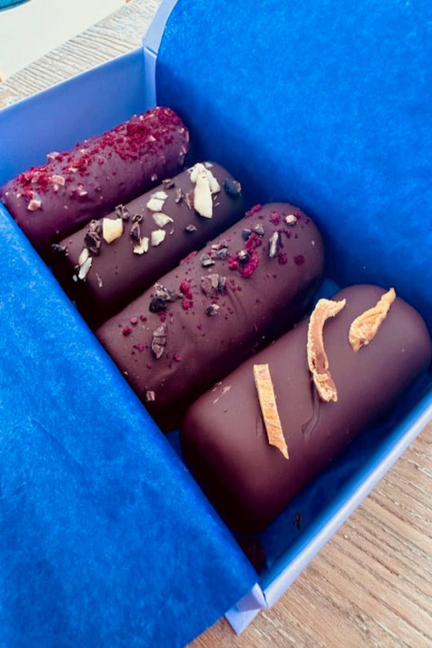 Four dark chocolate dessert logs, topped with dried mango, raspberries, and nuts, in a blue gift box.