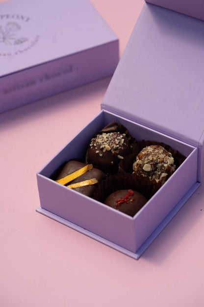 Purple box of four dark chocolate bonbons: two round truffles with toppings, two heart-shaped with orange zest.