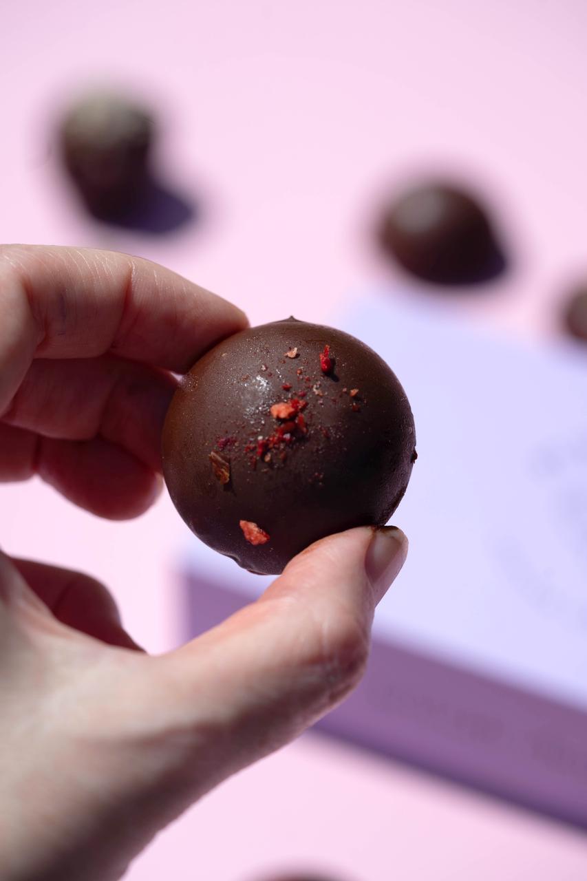 Hand holding a dark chocolate salted caramel cashew praline, sprinkled with red flakes, against a pink background.