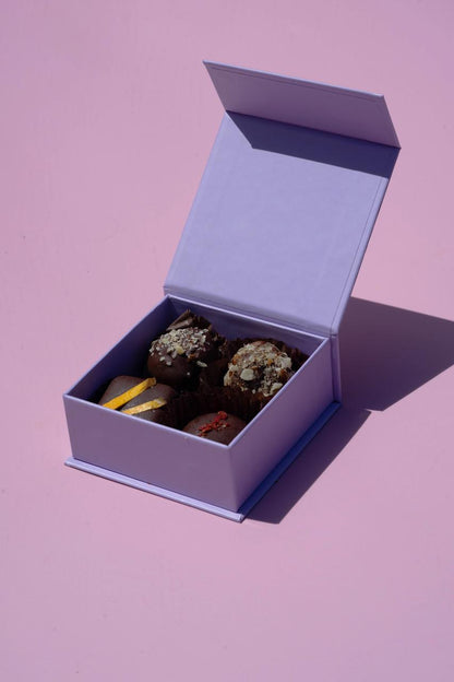 Open lavender gift box with six artisanal salted caramel cashew pralines, some topped with nuts or zest.
