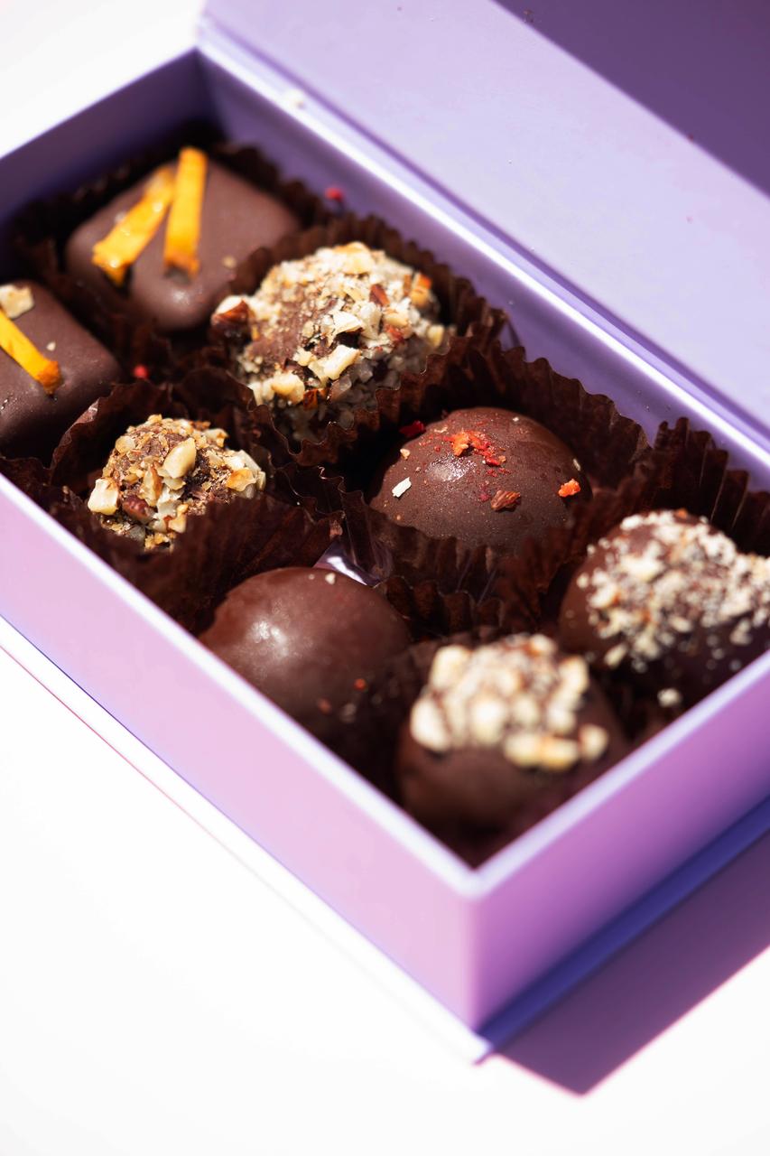 Open lavender box of assorted dark chocolate salted caramel cashew pralines, some topped with nuts or orange peel.