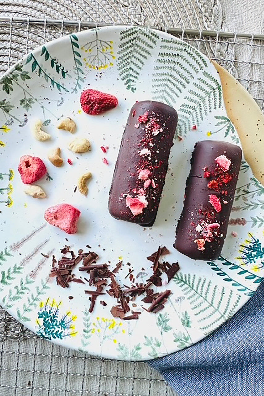 Two dark chocolate salted caramel dessert logs topped with pink sprinkles, on a botanical plate with cashews and dried raspberries.