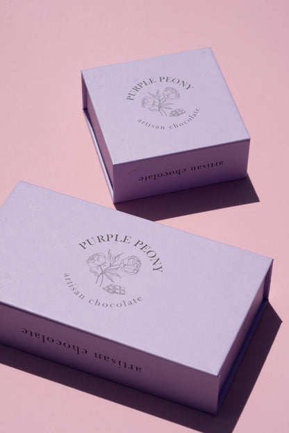 Two light purple "Purple Peony Artisan Chocolate" boxes with floral and chocolate bar designs on a pink background.